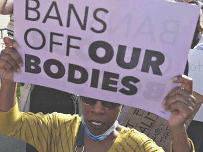 A black woman holding a purple sign that says "bans off our bodies" as a means to call out the white supremacy ideology