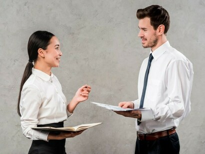 Two young professionals discuss a project against a grey wall — a scene that hides the often unseen cost of getting in