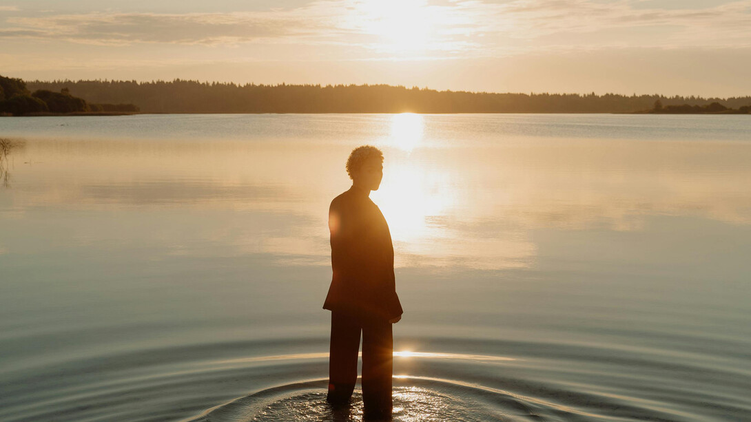 A figure stands at the shore of a lake, illuminated by the sun. 