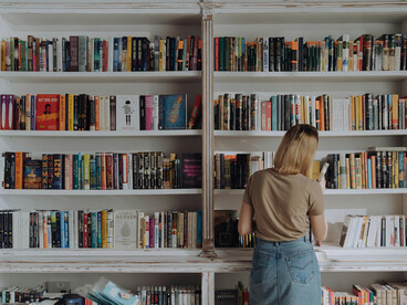 A woman in a beige long-sleeve shirt stands in front of a white wooden bookshelf filled with books