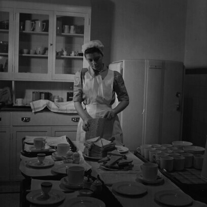 Estudiante de enfermería corta pan para sus pacientes en la cocina de la sala del Hospital St Helier. Está preparando té, huevos duros y pan con mantequilla. 1943