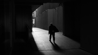 Una figura solitaria avanza por un pasaje oscuro hacia una zona iluminada, dejando una larga sombra sobre el suelo