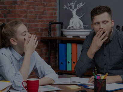 Young female and male co-workers sitting at office table, yawning from exhaustion, highlighting workplace challenges and the need for improvement