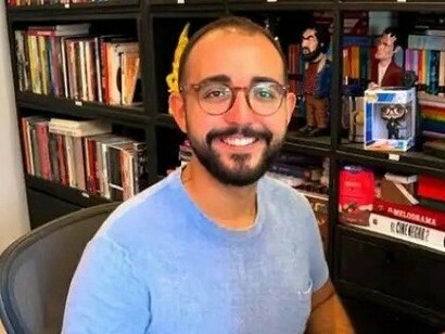 Raphael Montes posa diante de uma estante repleta de livros, em um ambiente que evidencia sua relação com a literatura. O escritor construiu sua trajetória explorando o suspense, o crime e os aspectos mais sombrios da psique humana