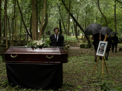 A brown coffin surrounded by mourners dressed in black during a forest funeral