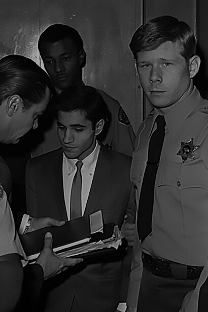 Sirhan Bishara Sirhan, ringed by deputy sheriffs, is handed documents by defense investigator Michael A. McCowan on way to courtroom, 1969