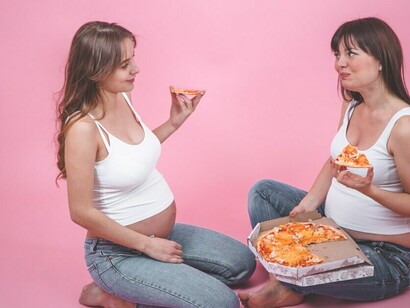 Pregnant women enjoying pizza in front of a pink wall, highlighting the impact of Western diet foods and processed foods on health risks during pregnancy