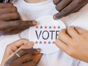 Mãos de diferentes pessoas seguram um adesivo relacionado ao voto diante de uma jovem sorridente. A democracia se constrói justamente pela participação de indivíduos diferentes em uma mesma esfera de escolha