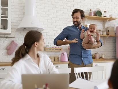 In a sunlit kitchen, a father soothes his crying baby while his partner types away at her laptop, celebrating the flexibility to care for family and work in the same space despite occasional interruptions