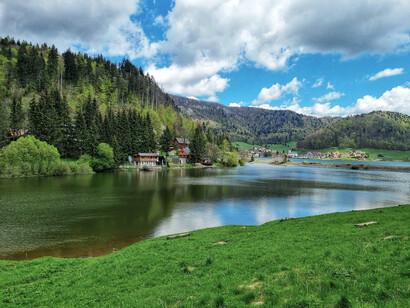 Green trees line the lake under a blue daytime sky, with Palcmanská Maša and the village of Dedinky in the background, Slovakia