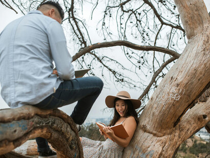 Hombre con camisa de vestir blanca y jeans azules sentado en la rama de un árbol leyendo un libro junto a una mujer con vestido floral blanco y negro y con un sombrero marrón, quien también está leyendo un libro