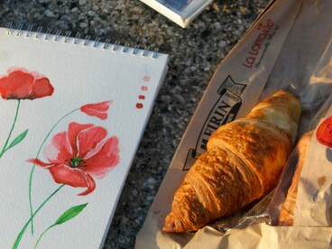 Um bloco de aquarelas, uma caixa de tintas e um croissant compõem um cenário de criação artística ao ar livre. Com cafés, esboços e pequenas pausas, François transforma as impressões da viagem em futuras pinturas