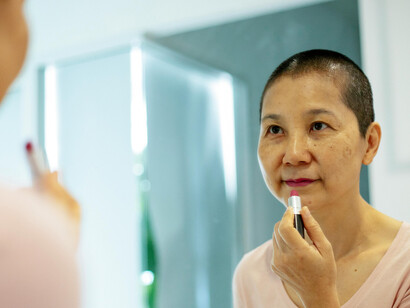 An Asian woman undergoing breast cancer treatment stands near a mirror, applying lipstick