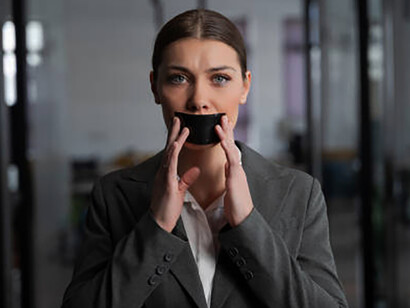 A woman in a gray suit stands in a modern office, her mouth covered with black tape, looking concerned, symbolizing political persecution under an authoritarian regime