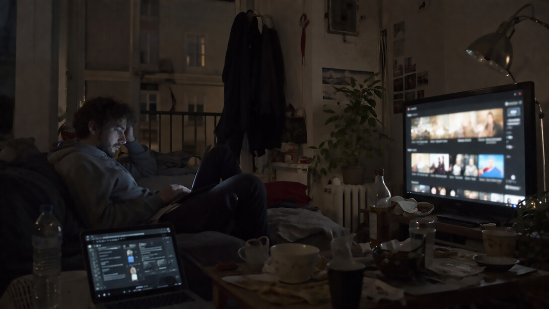 A young man sits alone in a dimly lit room, surrounded by screens, absorbed in a digital world