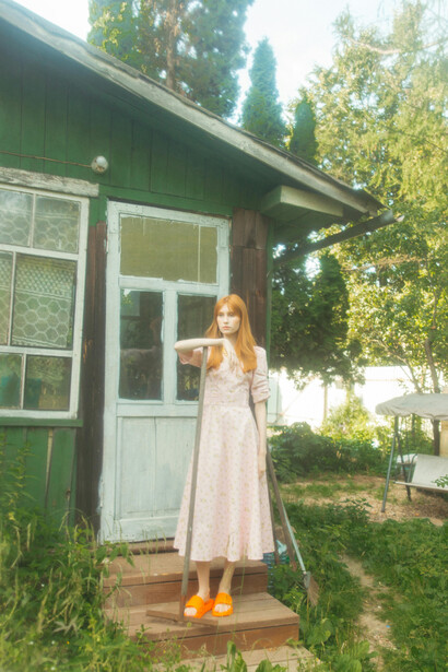 Bathed in warm summer light, a young woman in a vintage dress stands at the doorway of a rustic country house, framed by vibrant greenery and flowers