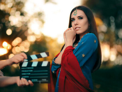 A stunning Bollywood actress in an ornate Indian ensemble with gold jewelry, performing on a grand film set while a dedicated crew captures the magic of cinema against a picturesque backdrop