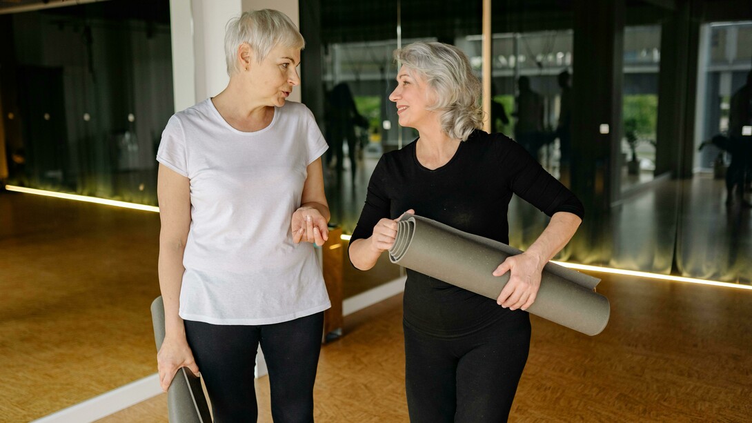 Two senior women chatting and smiling while holding yoga mats inside a modern fitness studio