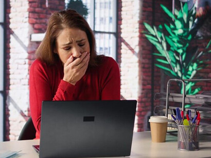 A burned-out woman sits at her loft desk, staring at her laptop and yawning from exhaustion