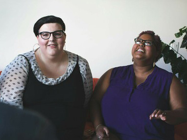 Two plus-size women are sitting on the couch, engaged in conversation while looking at each other
