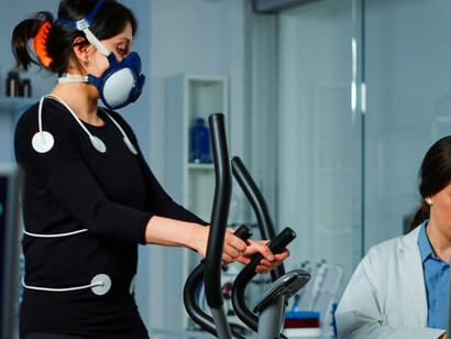 A medical researcher using a laptop to measure a sportswoman's endurance, with body sensors, electrodes, and a mask monitoring her cardiac rhythm