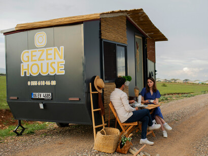A man and a woman seated on a brown wooden chair beside a black tiny portable house