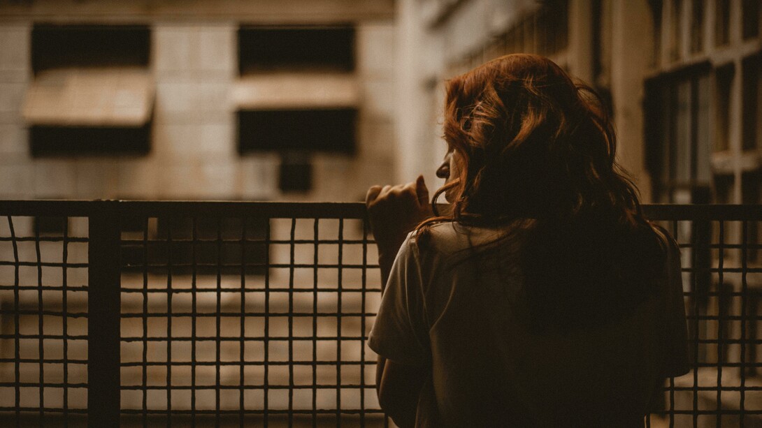 A woman stands alone on a balcony, gazing through the railing in a sepia-toned scene with a quiet and contemplative atmosphere