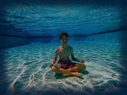 A boy sits on the bottom of the pool with his eyes open, legs crossed