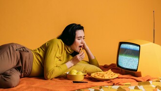 A woman in a yellow long-sleeve shirt watches TV while eating, binge-watching