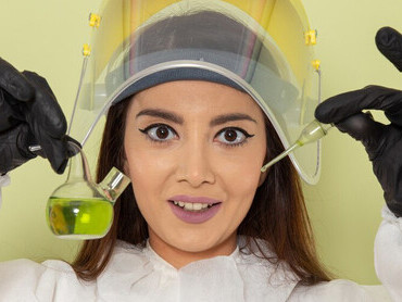 A female chemist in a protective suit holding a green solution on a green surface, carefully smelling it for analysis