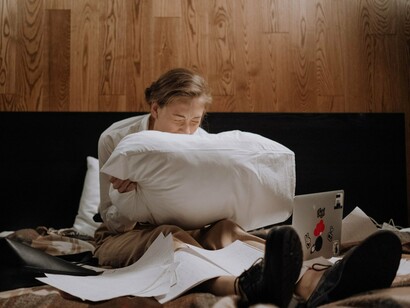 A woman screaming into a pillow while working from bed, representing the emotional weight of balancing productivity with chronic discomfort