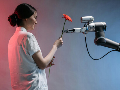 A robot holds a flower and gently offers it to a woman in a white T-shirt