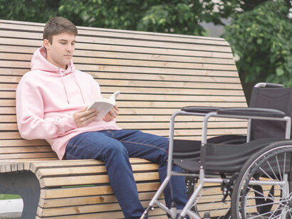 A man sitting on a bench reading a book next to his wheelchair