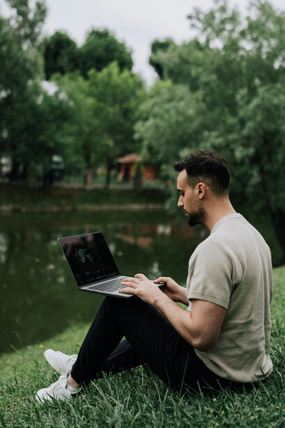A man relaxes on the grass beside a peaceful pond in a park, working on his laptop while enjoying the natural surroundings