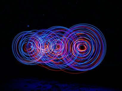 Feixes de luz formam círculos luminosos sobre um fundo escuro, criando uma atmosfera abstrata e hipnótica. Em meio ao ruído contemporâneo, o texto procura brechas de lucidez através da ironia e do humor