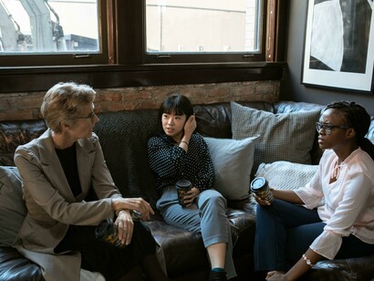 Three women, a Black woman, an Asian woman and a Caucasian woman stand together at work holding disposable cups and chatting