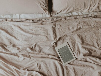 Um leitor de livros digitais repousa sobre os lençóis claros de uma cama cuidadosamente arrumada, transformando a tecnologia em objeto de leitura e descanso. Entre o livro físico e a tela, permanece a mesma busca pela atenção de quem lê