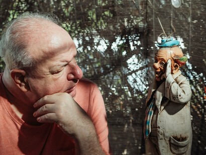 A fotografia mostra um homem idoso observando atentamente uma pequena estatueta de palhaço antiga, em um ambiente de aparência doméstica e cercado por objetos. O gesto contemplativo sugere um encontro entre memória e identidade, remetendo à investigação interior proposta pelo texto e à busca por aquilo que permanece oculto sob as nossas defesas