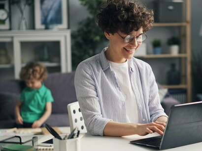 A mother at her workspace with her child playing behind her, reflecting the quiet overlap of professional and personal worlds