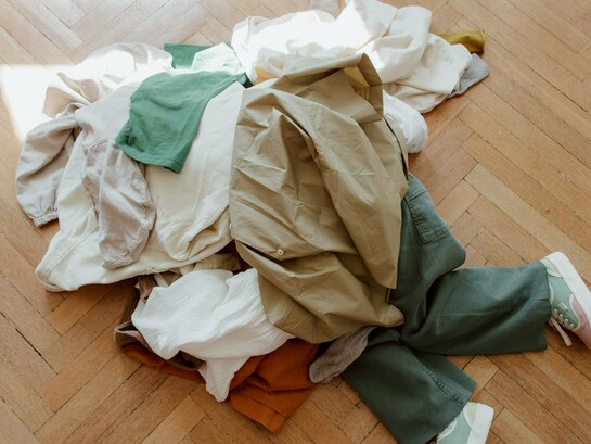 Diversas peças de roupa cobrem completamente uma pessoa deitada no chão. A imagem simboliza o excesso de consumo e convida à reflexão sobre a quantidade de vestuário adquirida e descartada atualmente. O cenário representa o impacto da cultura do consumo acelerado e a necessidade de escolhas mais conscientes