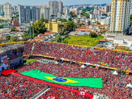 Na Vila Euclides, em São Bernardo do Campo, Lula retorna ao lugar que marcou o início de sua trajetória política para lançar sua campanha à reeleição. Diante de uma multidão que ocupa o estádio, uma gigantesca bandeira brasileira se estende pelo campo, enquanto a cidade se abre ao fundo. A imagem condensa décadas de história política brasileira e coloca em primeiro plano a disputa por um país cuja democracia, como argumenta Boaventura de Sousa Santos, volta a estar no centro de uma escolha que ultrapassa os limites de uma eleição