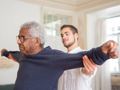 Um homem mais jovem, vestido de branco, segura os braços de um homem idoso durante um exercício de movimento. A luz natural que ilumina o ambiente reforça a atmosfera de confiança e acolhimento. A cena revela que o cuidado não se limita a atender às necessidades físicas: ele também preserva a proximidade, a dignidade e os vínculos humanos quando o corpo passa a exigir mais atenção