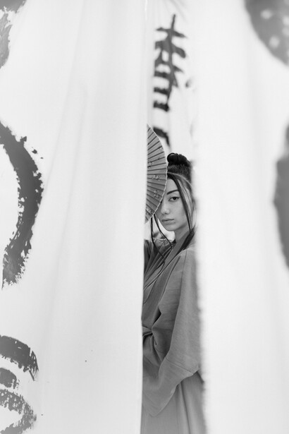 Joven japonesa sosteniendo un abanico frente a su rostro, oculta tras unas telas con caracteres escritos