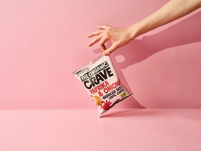 A person holding a pink and white plastic package of organic lentil-based chips from The Organic Crave Company
