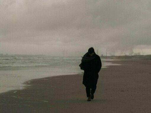 A solitary man walking along a desolate shoreline, representing the weight of regret and the longing to return home