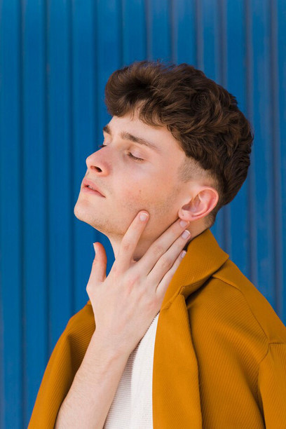 Trendy young man with a well-defined jawline standing confidently against a vibrant blue wall, wearing contemporary clothing that highlights his facial structure