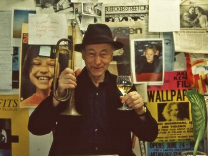 Jonas Mekas holding a glass of wine in one hand and a trumpet in the other