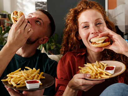 Young woman glancing at her boyfriend’s fast-food meal, highlighting the temptation of ultra-processed foods and unhealthy eating habit