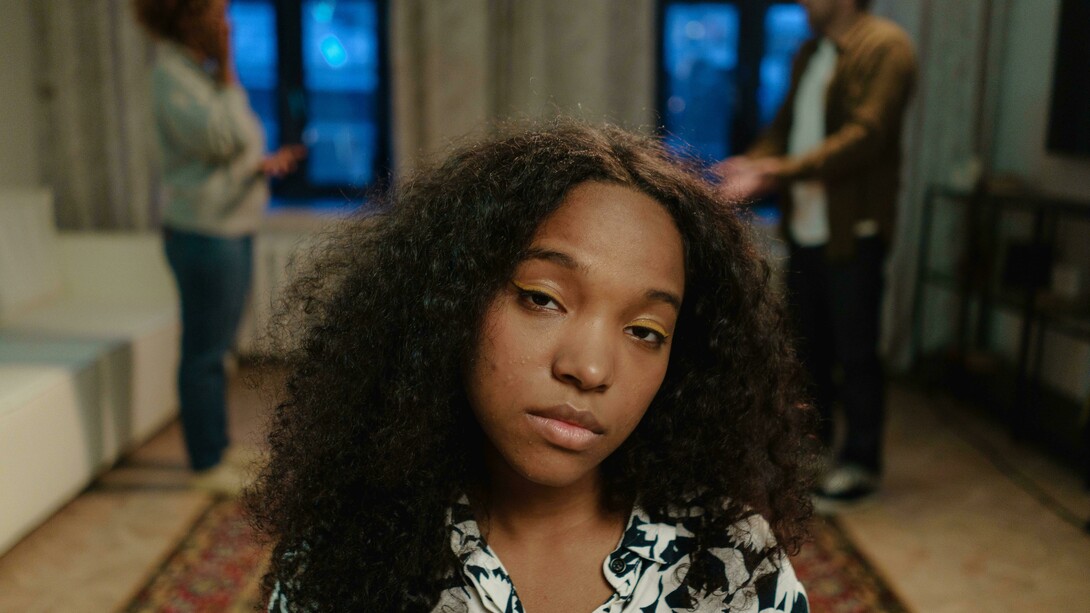 A concerned teenage girl with curly hair stands indoors while two adults argue in the blurred background