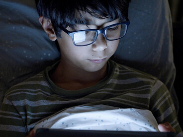 A boy wearing eyeglasses uses a digital tablet at night, focused on the screen